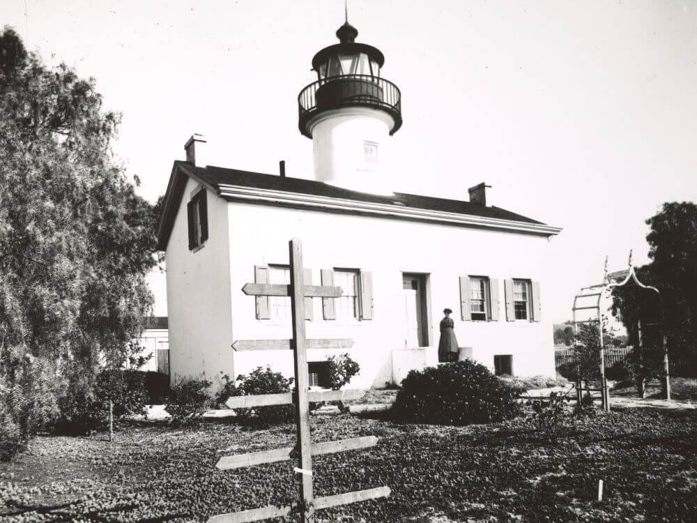 a woman stands next to the doorway of a long narrow building topped with a lighthouse