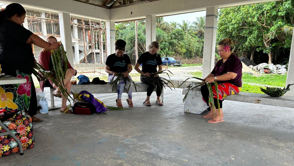 five people together under an outdoor pavilion weaving coconut fronds