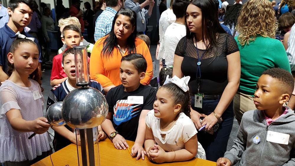 Young children gather around a Van de Graaff machine, a metal structure with a large metal sphere on top, with a metal spherical wand attached. The children watch the machine with faces of awe as they see electricity passing between the machine and the wand.