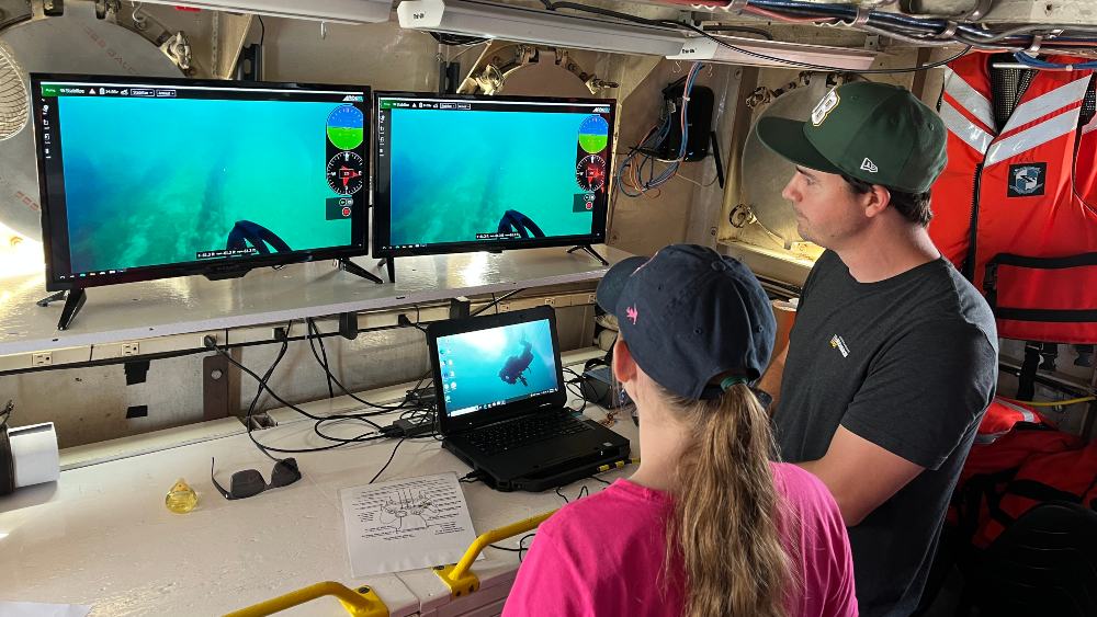 A young female student and a researcher look at several monitors showing them underwater footage of shipwrecks in Wisconsin Shipwreck Coast National Marine Sanctuary.