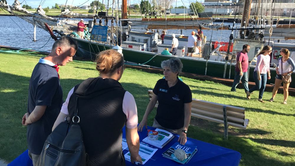 A person in a shirt with a NOAA logo staffs an outreach booth with educational handouts at a maritime festival.
