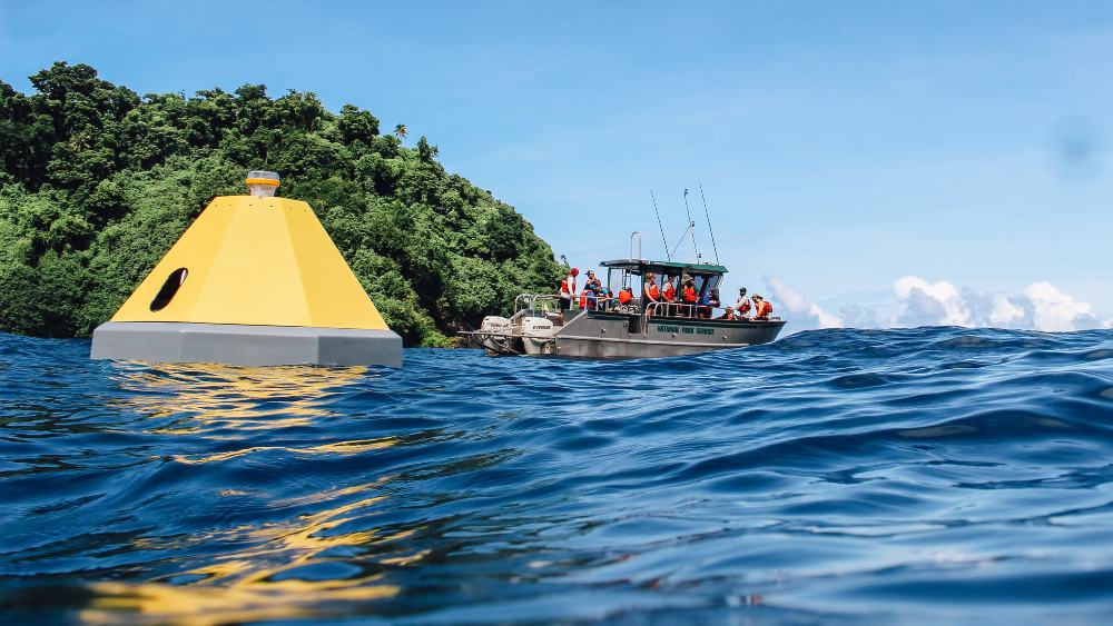 A yellow monitoring bouy sits at the top of the water in the foreground. A research boat full of people can be seen in the background.