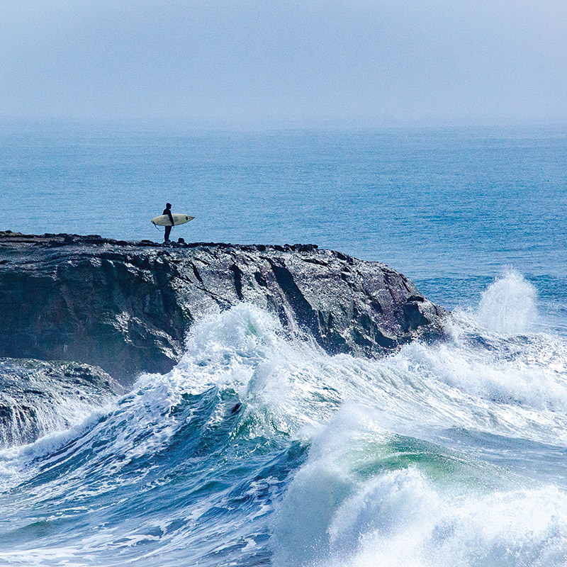 surfer stands at the rocky shore as wave crash by
