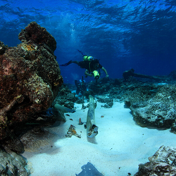 Two divers swim above metal debris on the seafloor