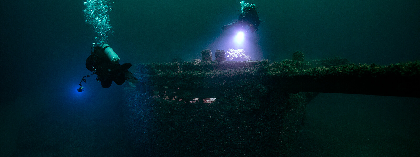 Two diver shine lights on shipwreck while they swim above it