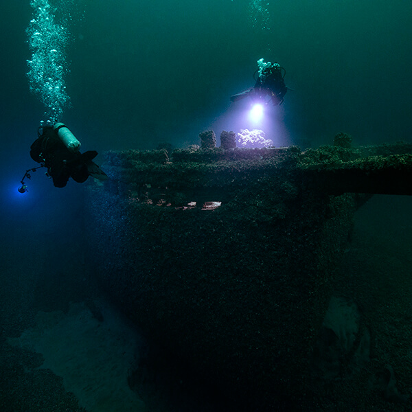Two divers shine lights on a shipwreck as they swim nearby