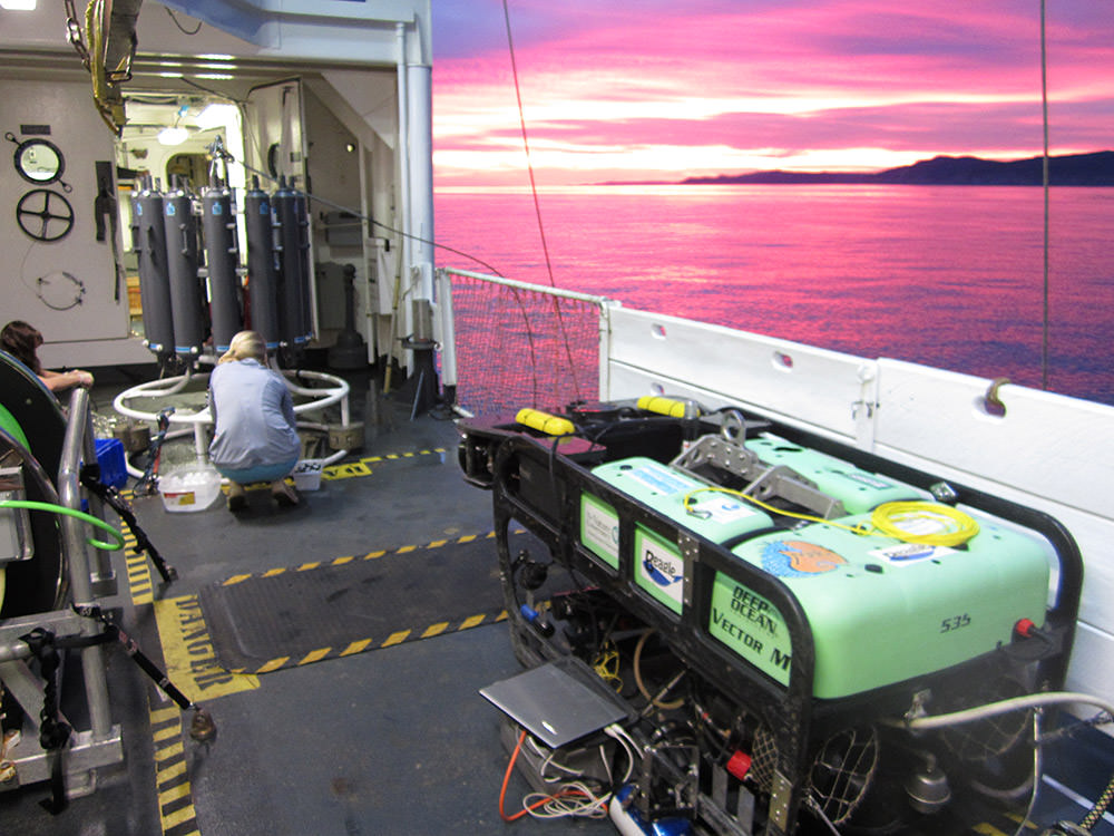sunset on the deck of the bell m. shimada, two scientist work on a water collection device next to the rov