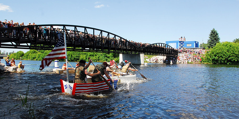 people paddling cardboard boat across a river while people watch on a bridge and on land